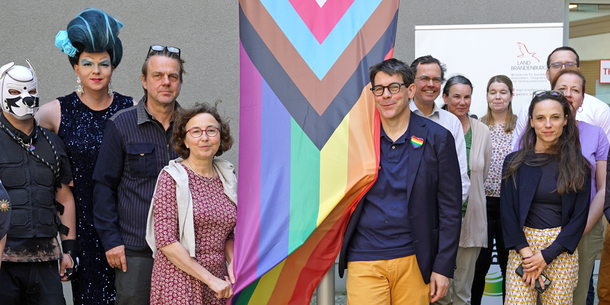 Das Foto zeigt mehrere Menschen, darunter Staatssekretär Götz und die Landesgleichstellungsbeauftragte Dörnenburg, beim Hissen der Regenbogenfahne vor dem Gebäude des Gleichstellungsministeriums in Potsdam.